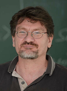 Herr Schaefer mit Brille, leichtem Bart und Poloshirt vor grüner Tafel.